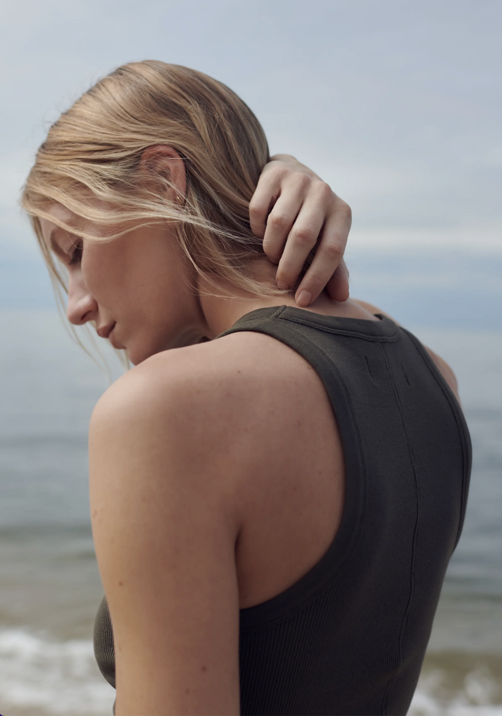 A blonde woman wearing The Not So Basic Tank by Sold Out NYC stands by the water, touching her neck and looking down thoughtfully against a calm, cloudy sky background.