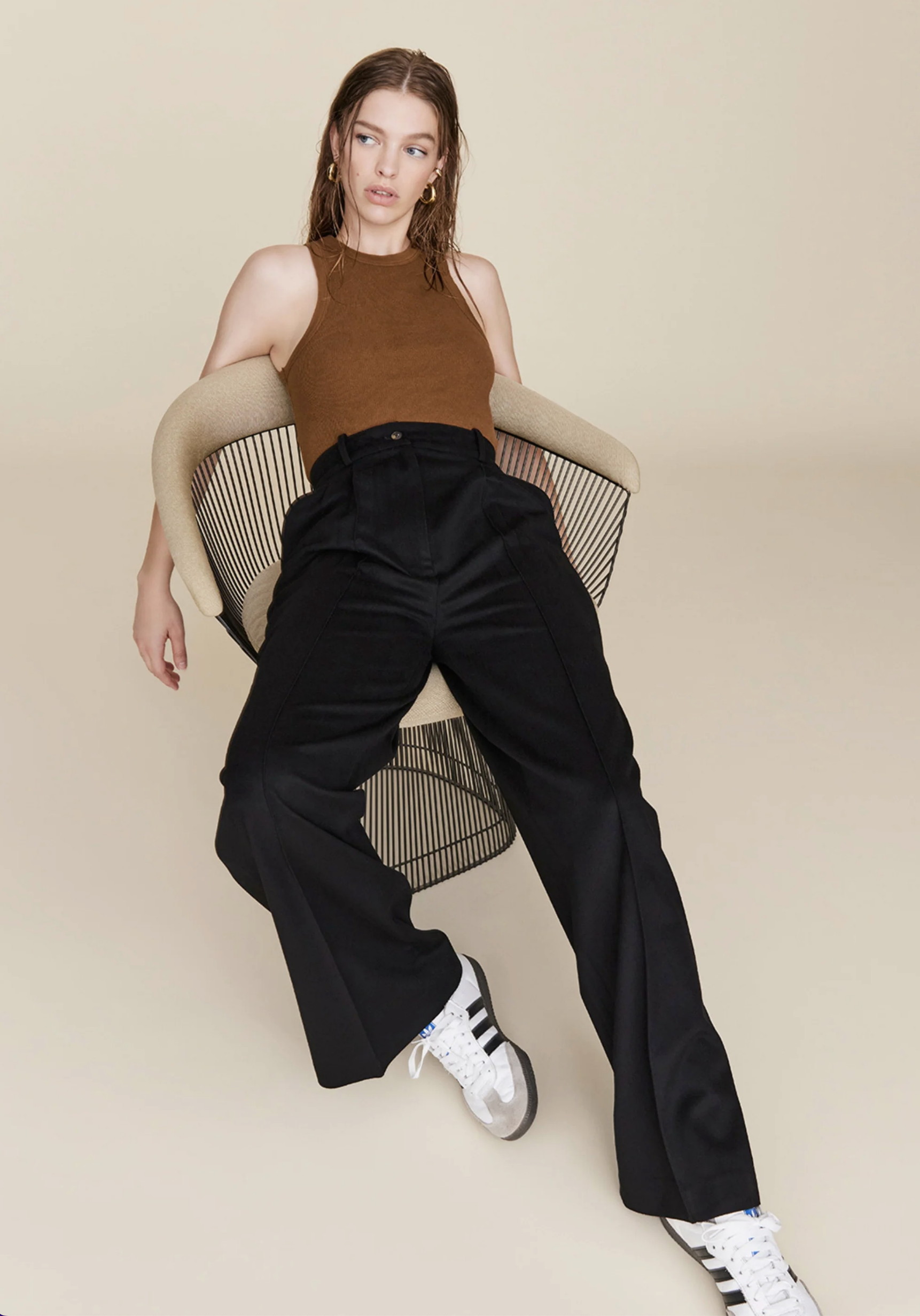 A woman with wet hair sits in a modern wire chair, wearing Sold Out NYC’s The Not So Basic Tank in brown, paired with high-waisted black pants and white Adidas sneakers, against a plain beige background.