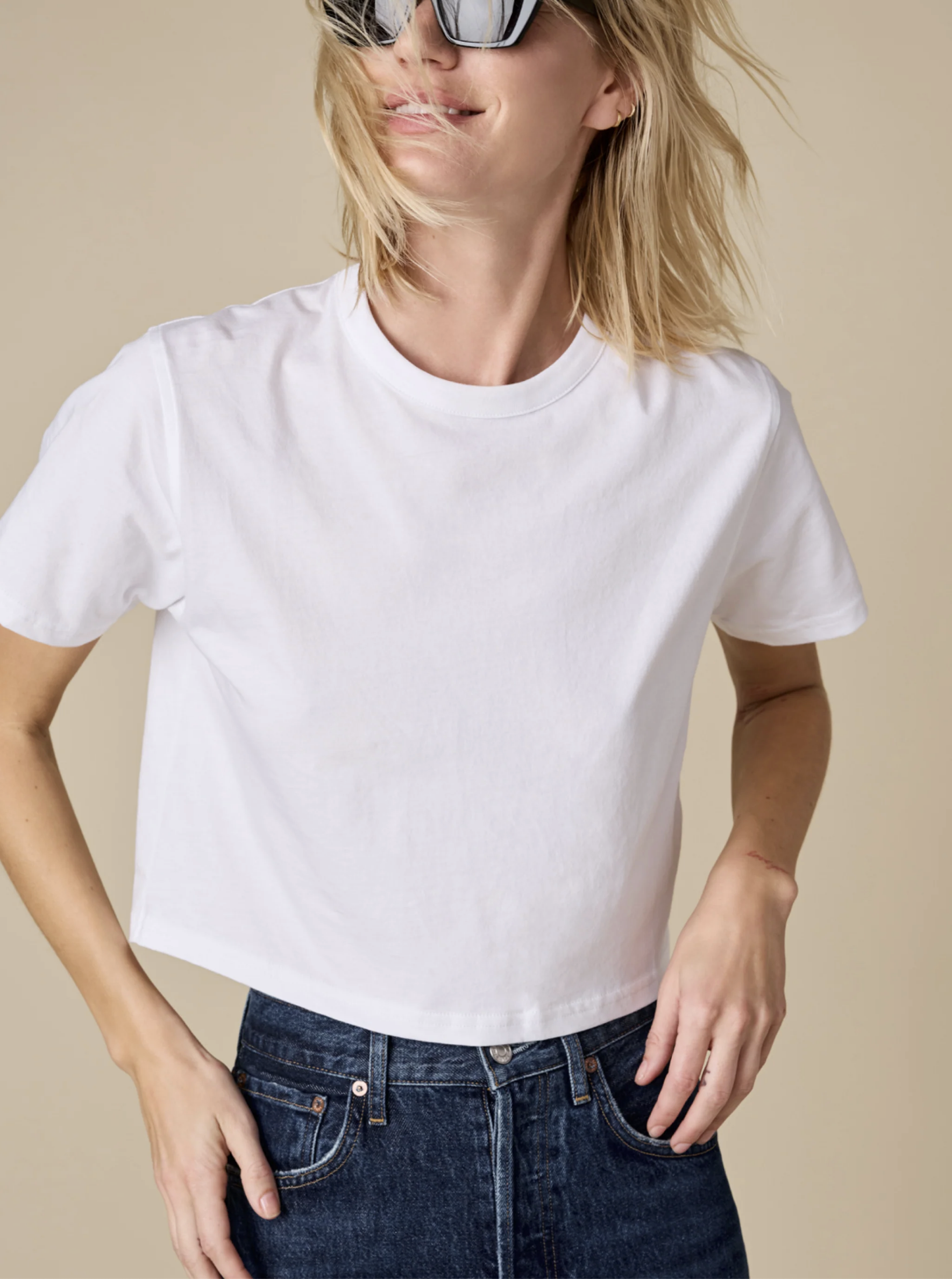 A woman in Sold Out NYC's The Cropped Perfect Tee, plain white Peruvian cotton, paired with dark blue high-waisted jeans and large sunglasses, smiles with tousled hair against a beige background.