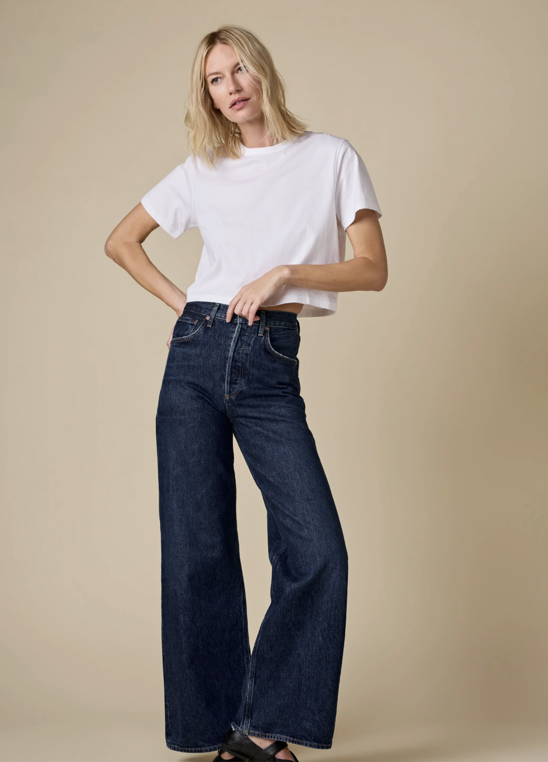 Wearing The Cropped Perfect Tee by Sold Out NYC and dark blue wide-leg jeans, a blonde-haired person stands before a beige backdrop, embodying the relaxed ease of a summer breeze.