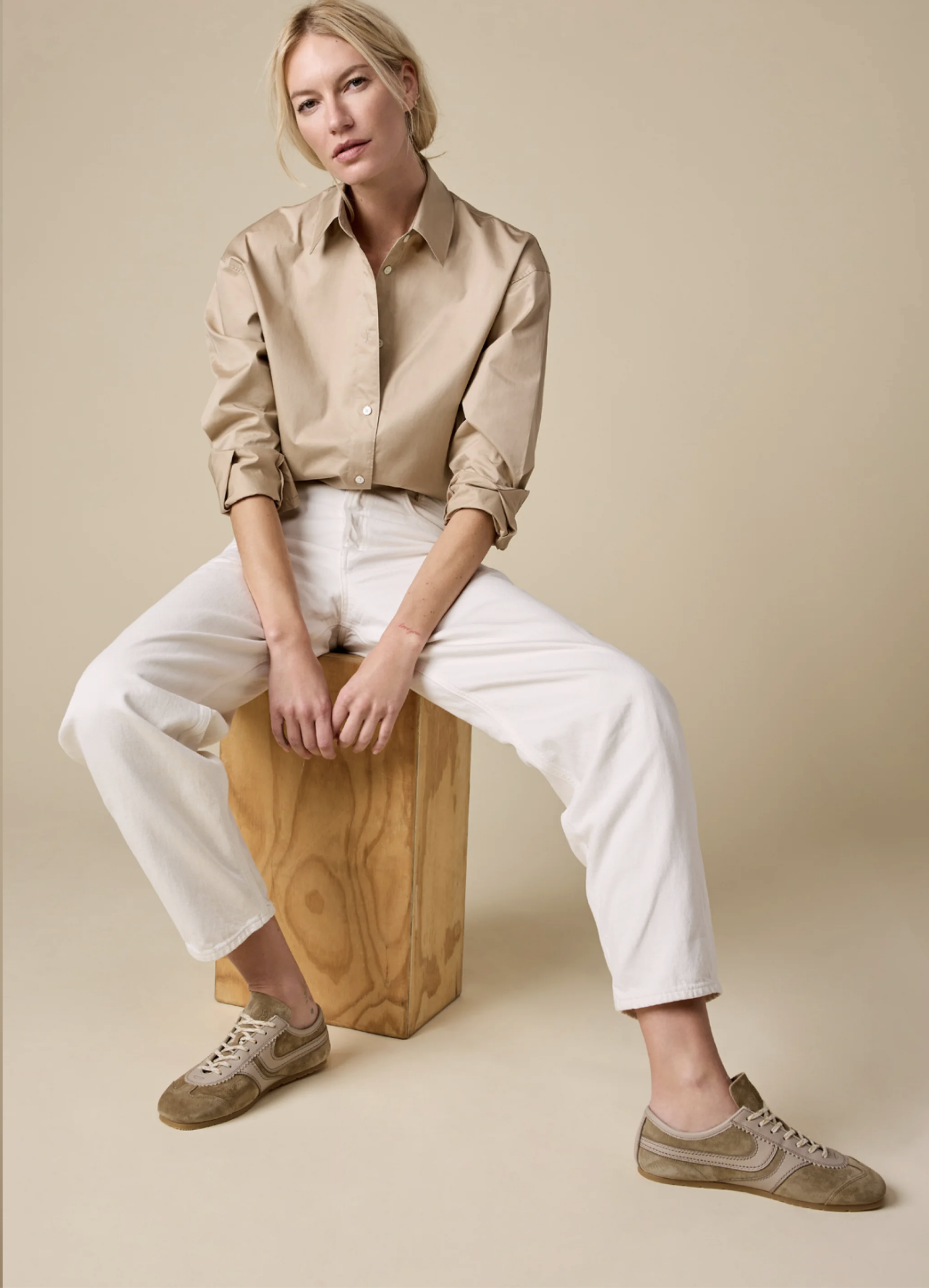 A person with blond hair sits on a wooden block against a beige background, wearing The Everything Shirt by Sold Out NYC—a 100% cotton tan button-up—paired with white pants and tan sneakers, arms resting on their knees and looking slightly to the side.