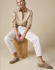 A person with blond hair sits on a wooden block against a beige background, wearing The Everything Shirt by Sold Out NYC—a 100% cotton tan button-up—paired with white pants and tan sneakers, arms resting on their knees and looking slightly to the side.