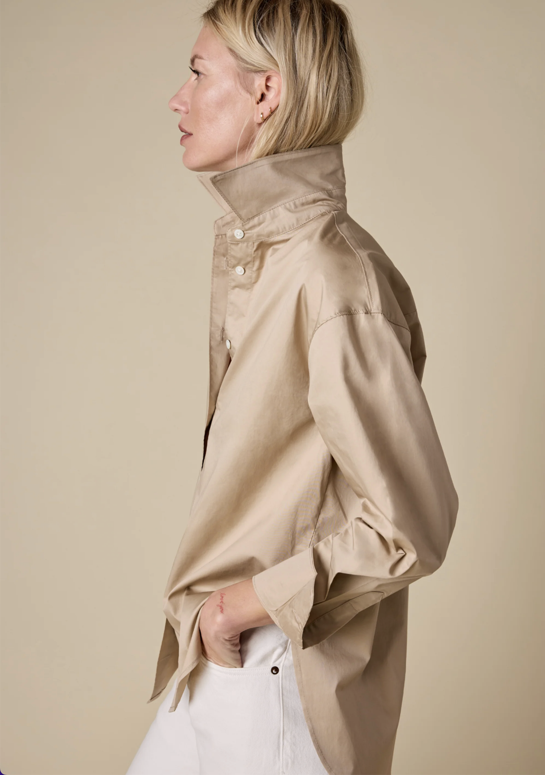 A person with blond hair stands in profile against a beige background, wearing Sold Out NYC’s The Everything Shirt—an oversized beige button-up with the collar popped—and white pants, with one hand in their pocket.