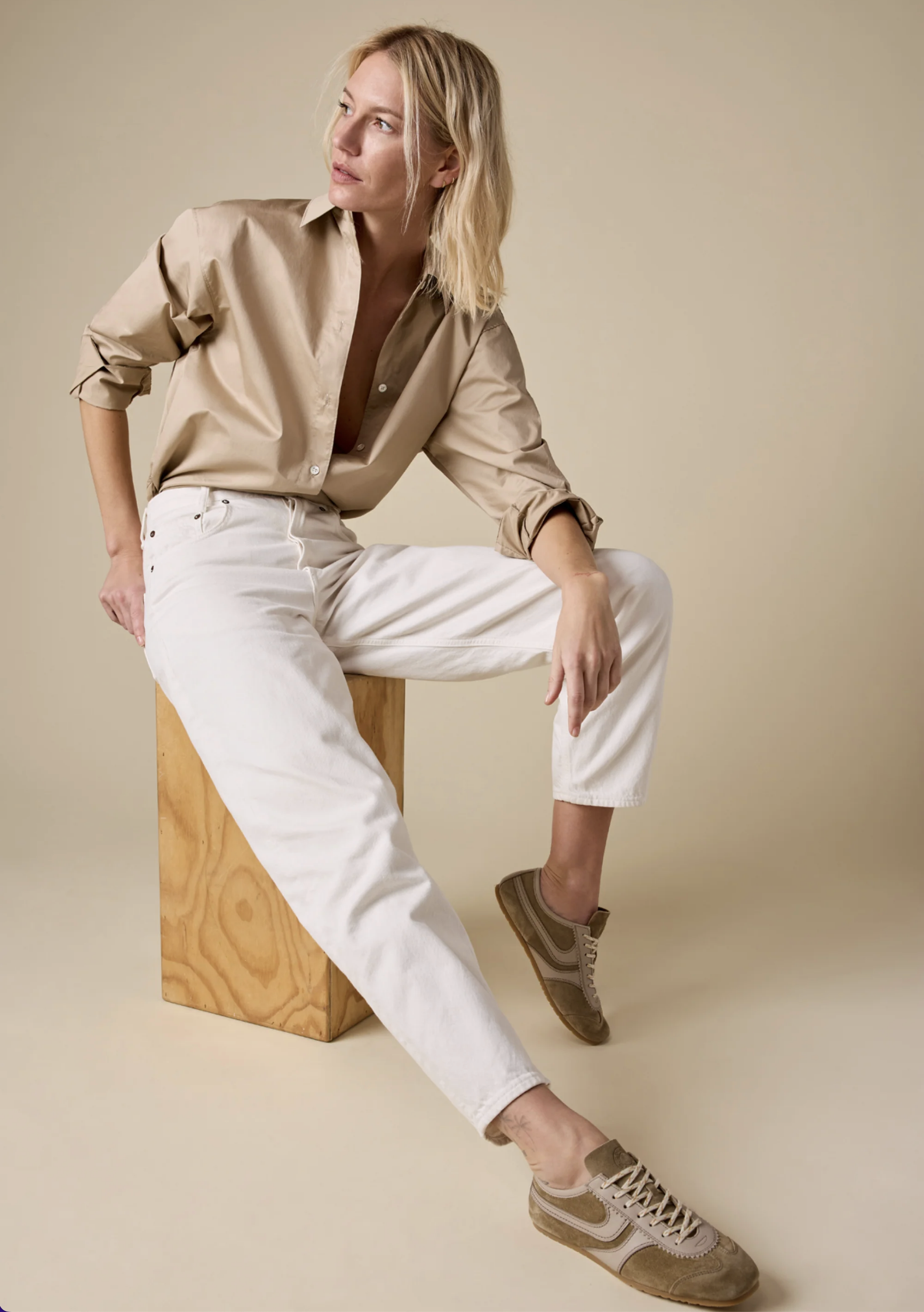 A person with blonde hair wears Sold Out NYC's The Everything Shirt, a 100% cotton beige button-up, paired with white pants and beige sneakers. Seated on a wooden block against a plain beige background, they look to the side.