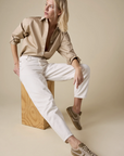 A person with blonde hair wears Sold Out NYC's The Everything Shirt, a 100% cotton beige button-up, paired with white pants and beige sneakers. Seated on a wooden block against a plain beige background, they look to the side.