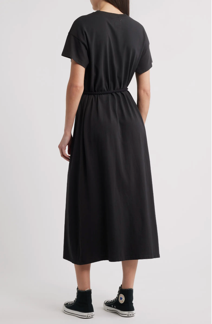 A person wearing The Great Inc.'s The Boxy Tee Dress—a short-sleeve, 100% cotton black midi dress with an elastic waist—and black high-top Converse stands facing away against a plain, light-colored background.