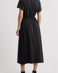 A person wearing The Great Inc.'s The Boxy Tee Dress—a short-sleeve, 100% cotton black midi dress with an elastic waist—and black high-top Converse stands facing away against a plain, light-colored background.