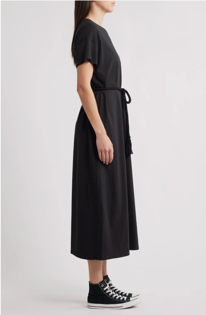 A woman stands in profile wearing The Boxy Tee Dress by The Great Inc., a 100% cotton, short-sleeve, belted T-shirt silhouette dress, paired with black high-top sneakers against a plain light background.