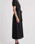 A woman stands in profile wearing The Boxy Tee Dress by The Great Inc., a 100% cotton, short-sleeve, belted T-shirt silhouette dress, paired with black high-top sneakers against a plain light background.
