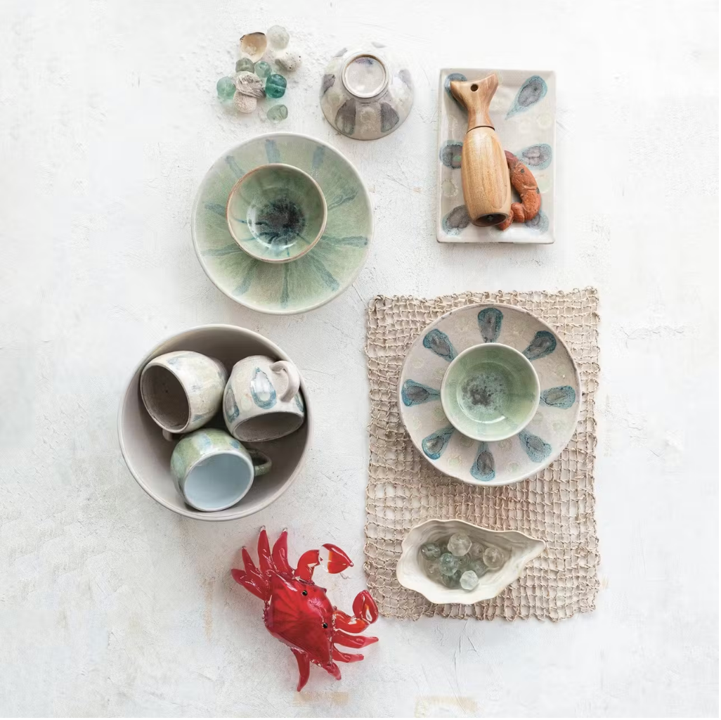 A flat lay features the Creative Co-op Low Bowl Blue Green with other ceramic dishes, cups, and bowls in blue-green patterns, a woven placemat, glass marbles, a wooden seal figurine, a red crab figurine, and a shell-shaped dish on white.