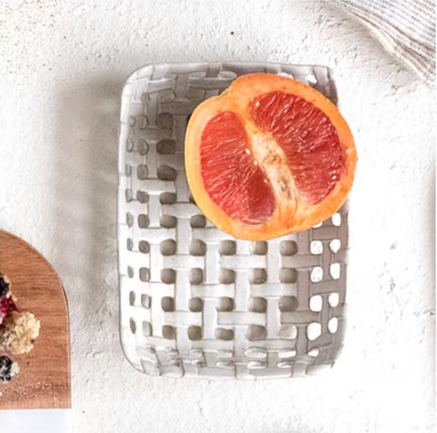 A halved red grapefruit rests on a postcard-sized Creative Co-op Square Lattice Dish atop a light surface. In the lower left, part of a wooden board with assorted fruits is visible.