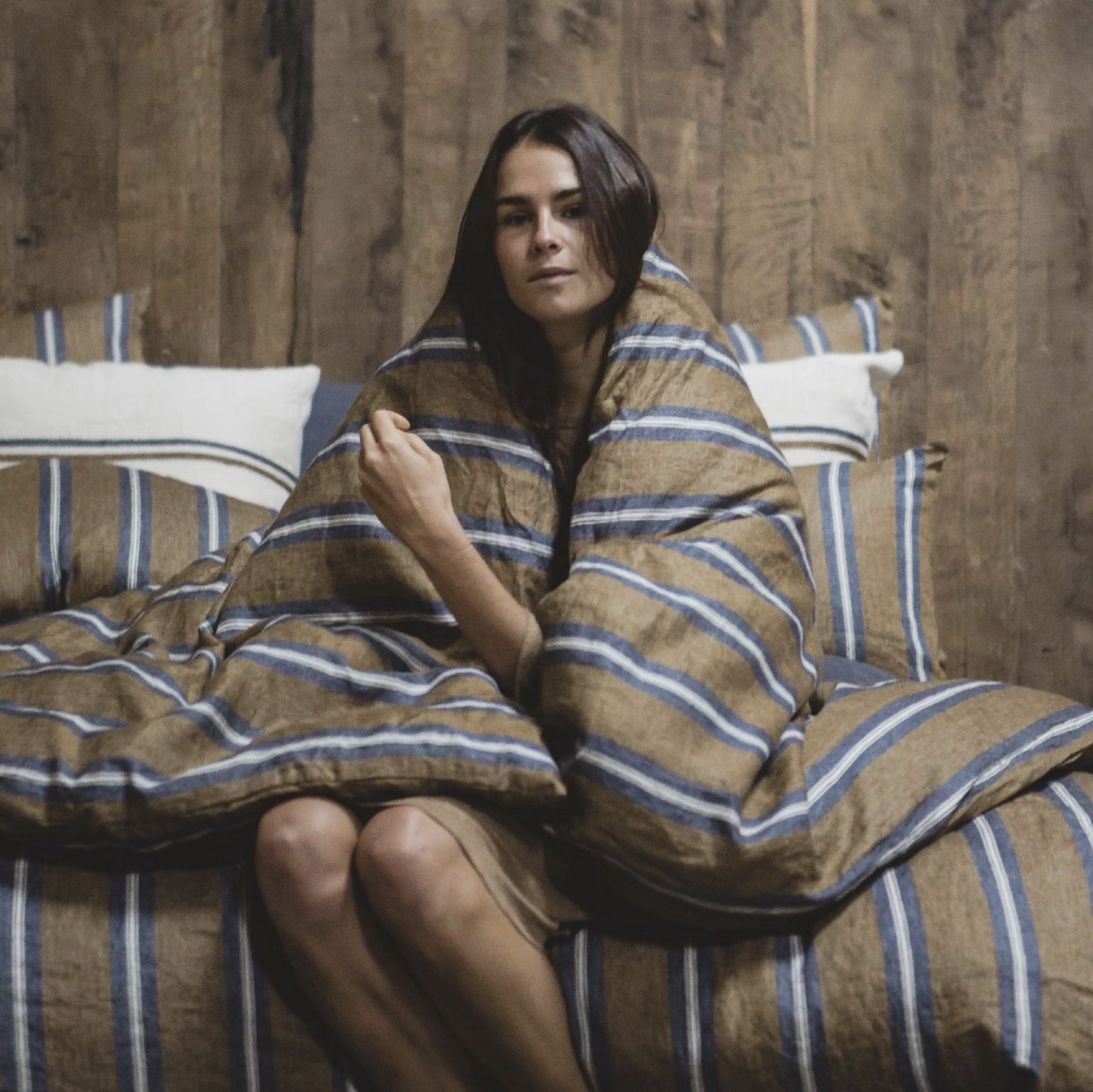 Wrapped in the Salem Duvet by Libeco Home, a woman sits on a bed with matching striped linen, gazing at the camera. The 100% linen fabric and wooden wall background create a warm, inviting atmosphere.