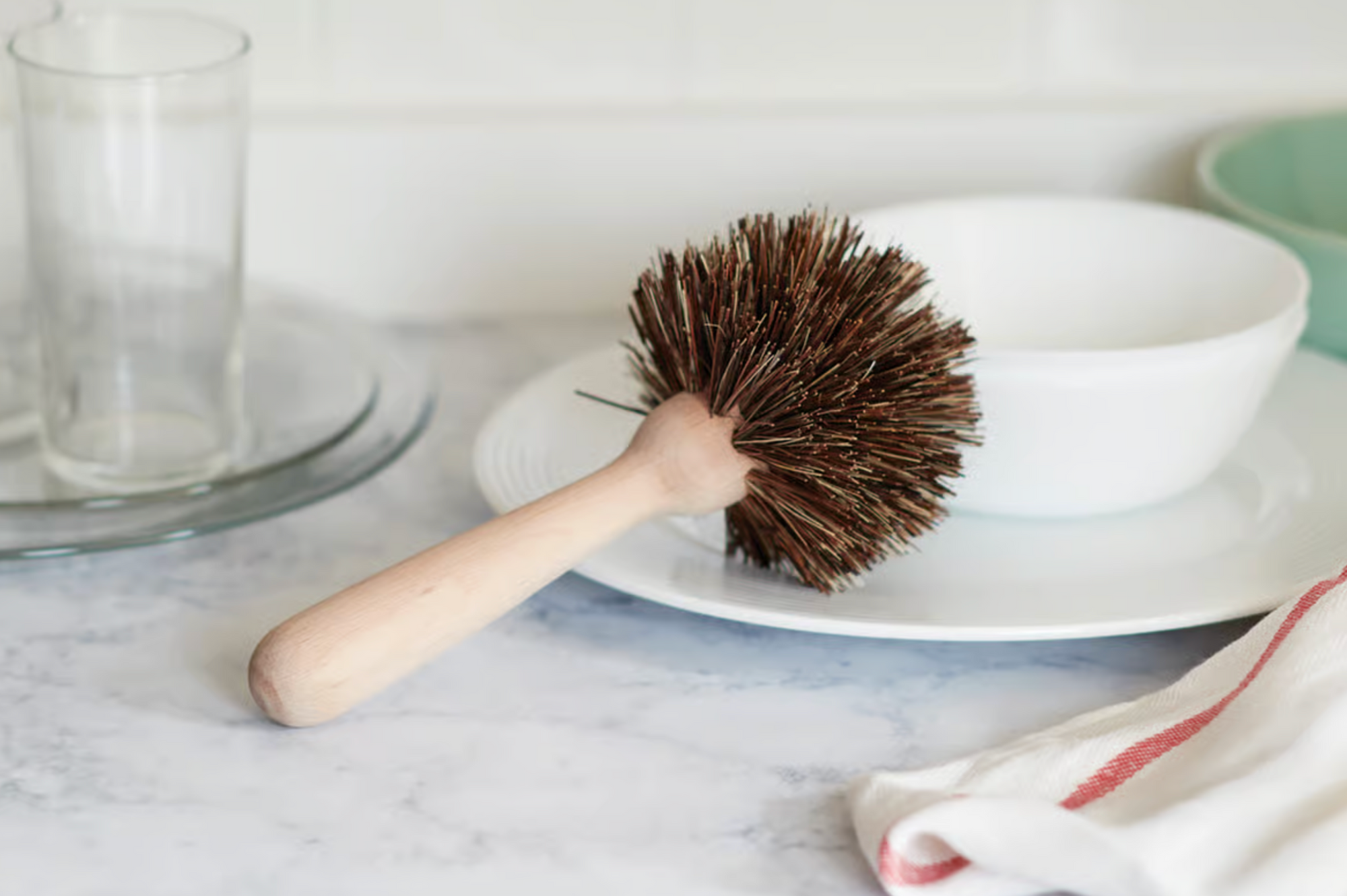 The Fox Run Brands Dish Brush, featuring natural bristles and a beechwood handle, sits on a white plate beside a bowl, glassware, and a red-striped dish towel on a kitchen countertop.