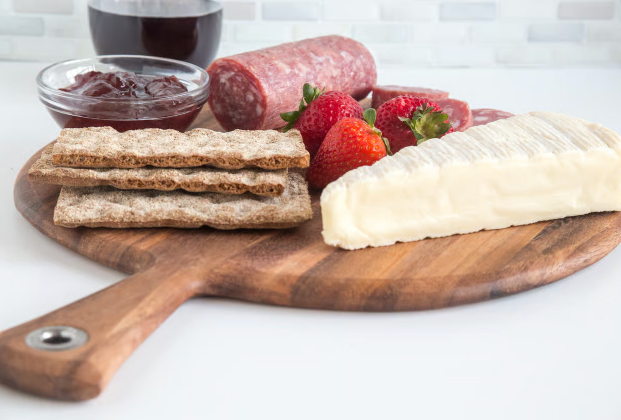 A beautifully arranged Provencale Acacia Board by Fox Run Brands holds sliced salami, crackers, brie cheese, strawberries, jam, and a glass of red wine for the perfect charcuterie spread.
