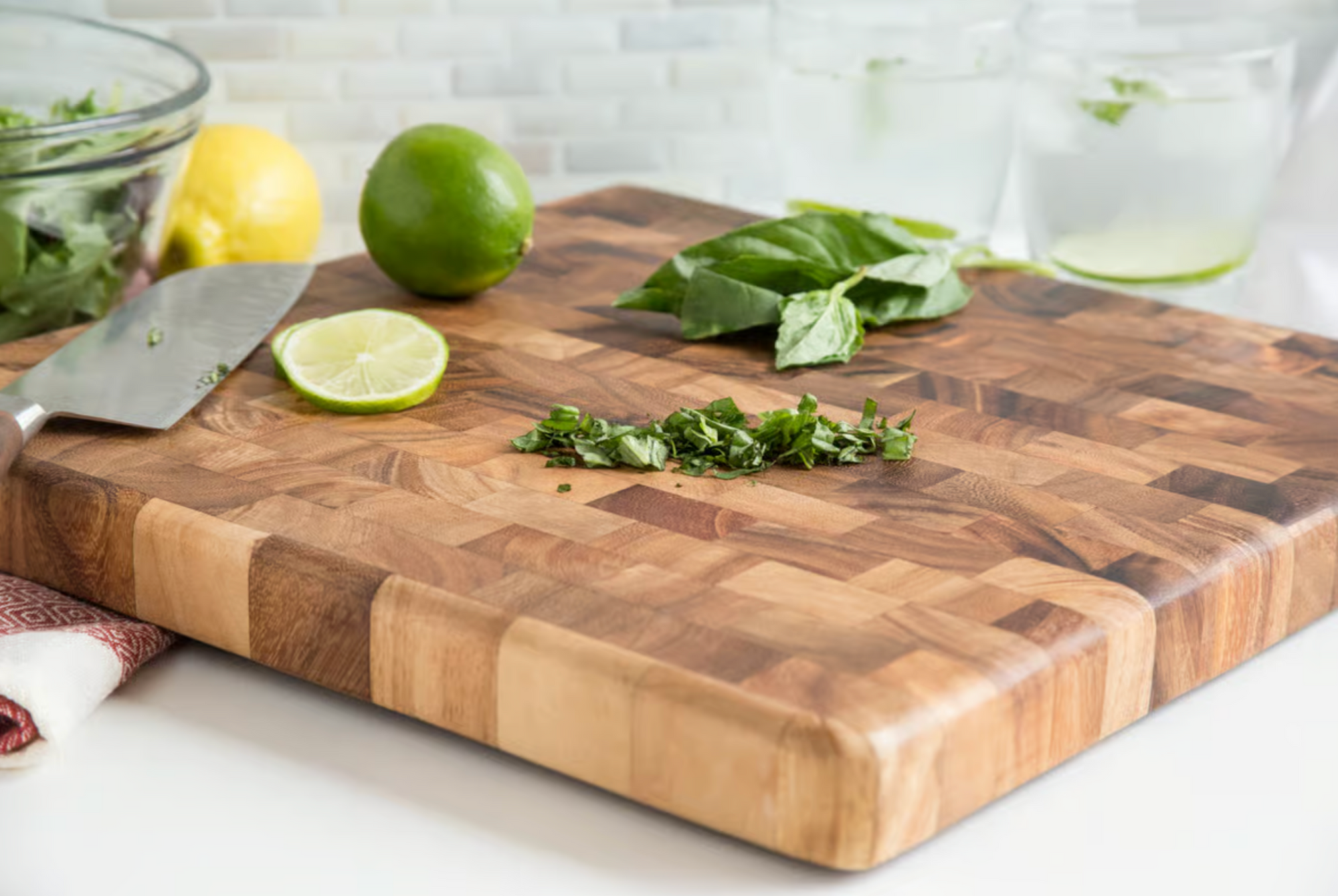 The Square Chef's Board by Fox Run Brands holds chopped herbs, a knife, basil leaves, a lemon, a whole and sliced lime. In the background are a bowl of greens and two glasses of water with lime.