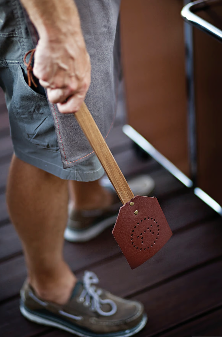 A person stands on a wooden floor holding the Fox Run Brands Acacia/Leather Fly Swatter, a heavy-duty brown leather swatter with a wooden handle.