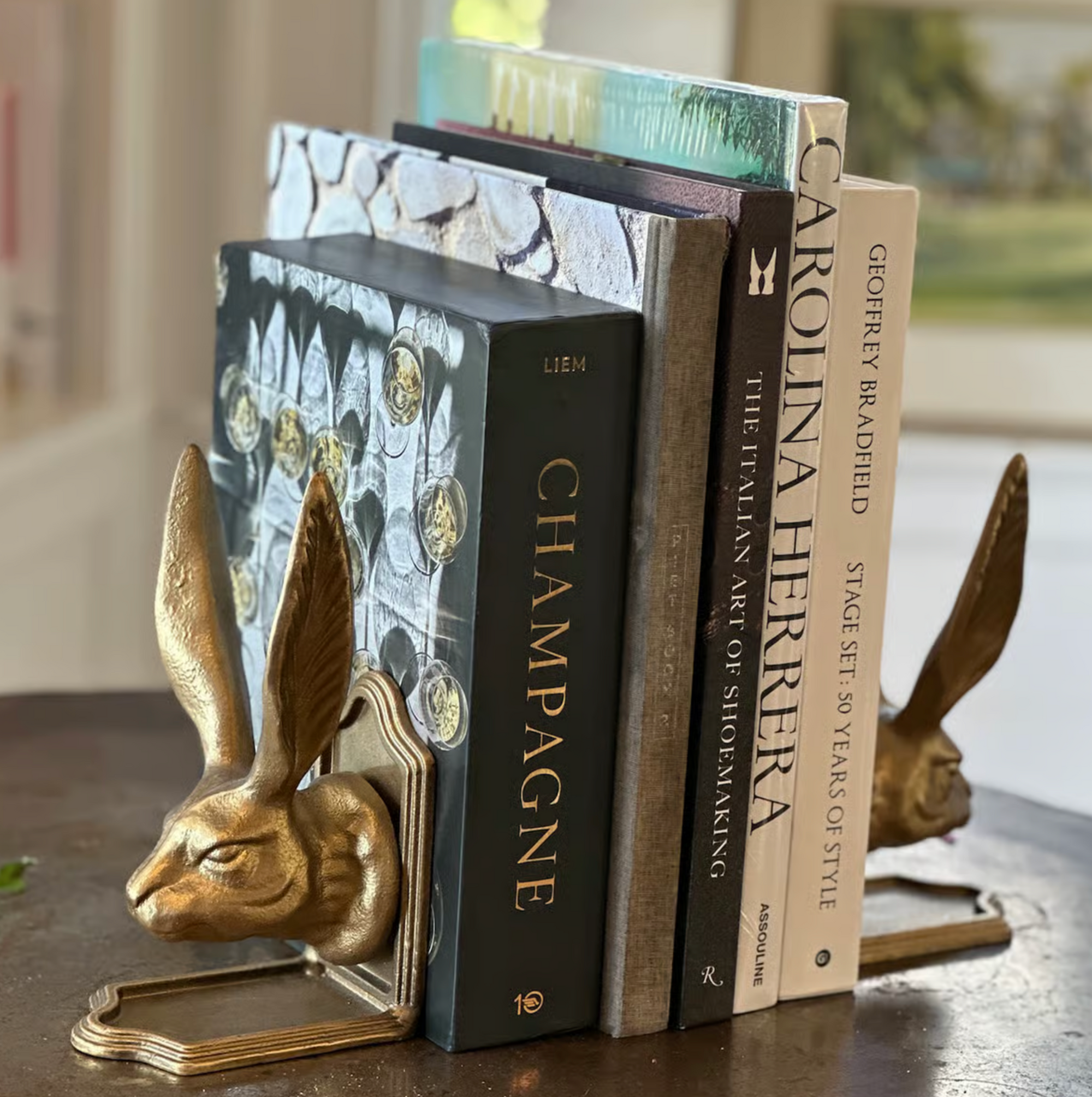 The Fox Run Brands Rabbit Bookend Set holds four books—"CHAMPAGNE," "The Italian Art of Shoemaking," "Carolina Herrera," and "Stage Set 50 Years of Style"—on a round wooden table.