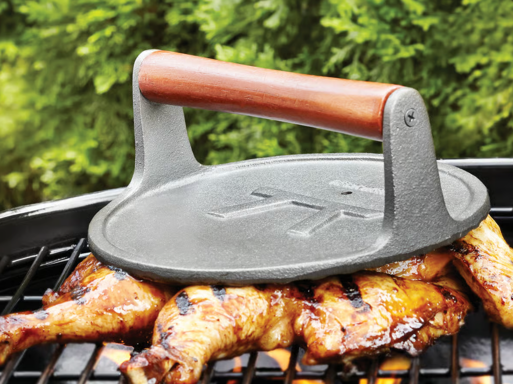 The Fox Run Brands Cast Iron Grill Press, featuring a wooden handle, rests atop grilled chicken on an outdoor barbecue and doubles as a bacon press, with green foliage visible in the background.