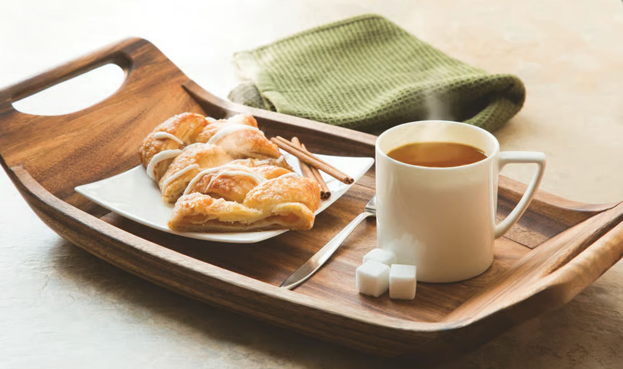 The Fox Run Brands Acacia Serving Tray holds a white mug of steaming tea, two sugar cubes, a plate with croissants and cinnamon sticks, with a folded green towel in the background.