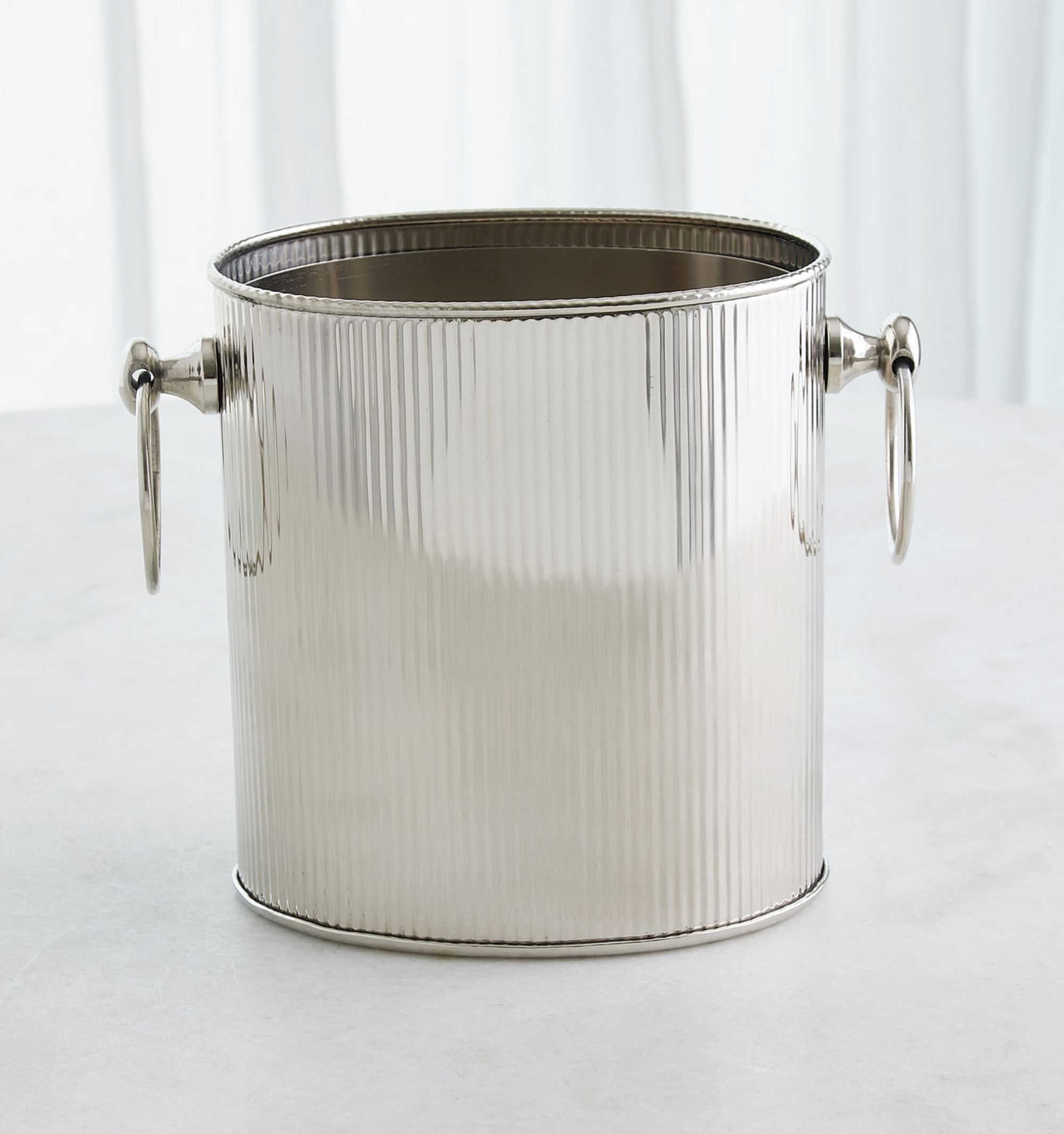 The Winston Ice Bucket L by Global Views, crafted from stainless steel with a fluted design and round side handles, rests on a light surface with a white curtain backdrop.