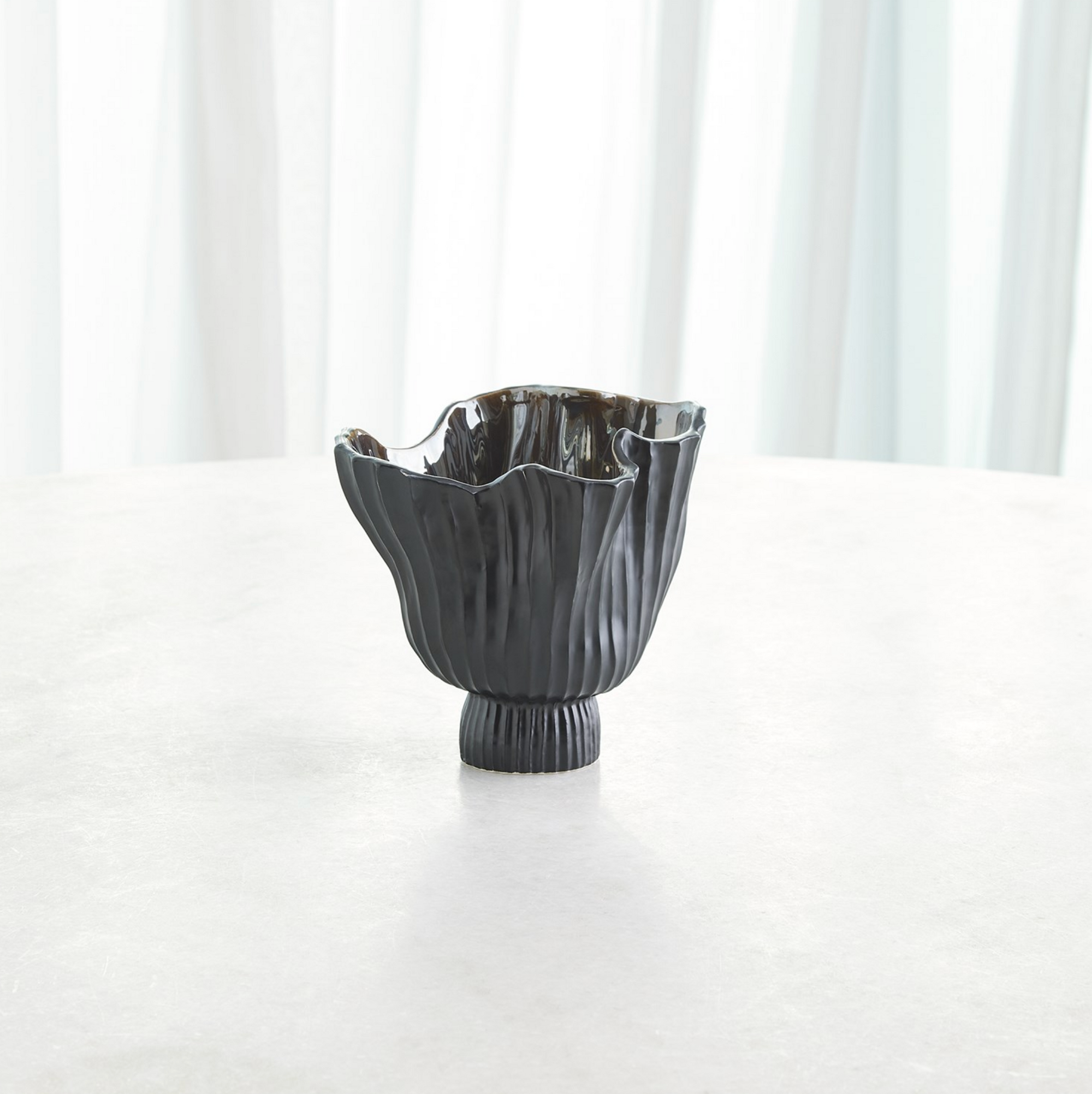 The Folha Footed Vase S by Global Views is a dark ceramic piece with a wavy, fluted rim and vertical ridges, hand-painted in metallic tones by Portuguese artisans. It sits on a round surface with sheer white curtains in the background.