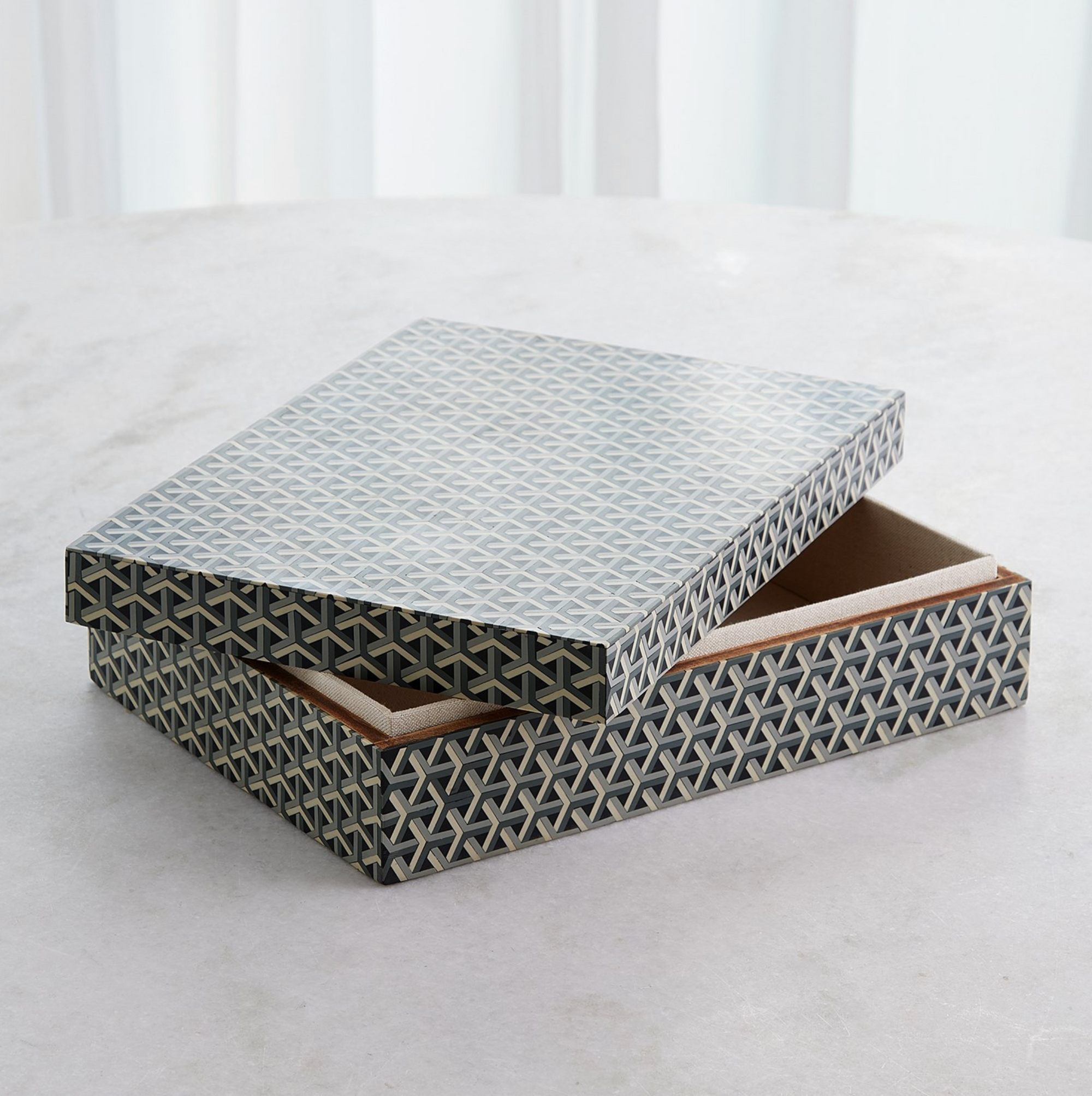The Interwoven Box LG by Global Views features a geometric grey pattern and is shown partially open on a light surface with a soft white curtain in the background.