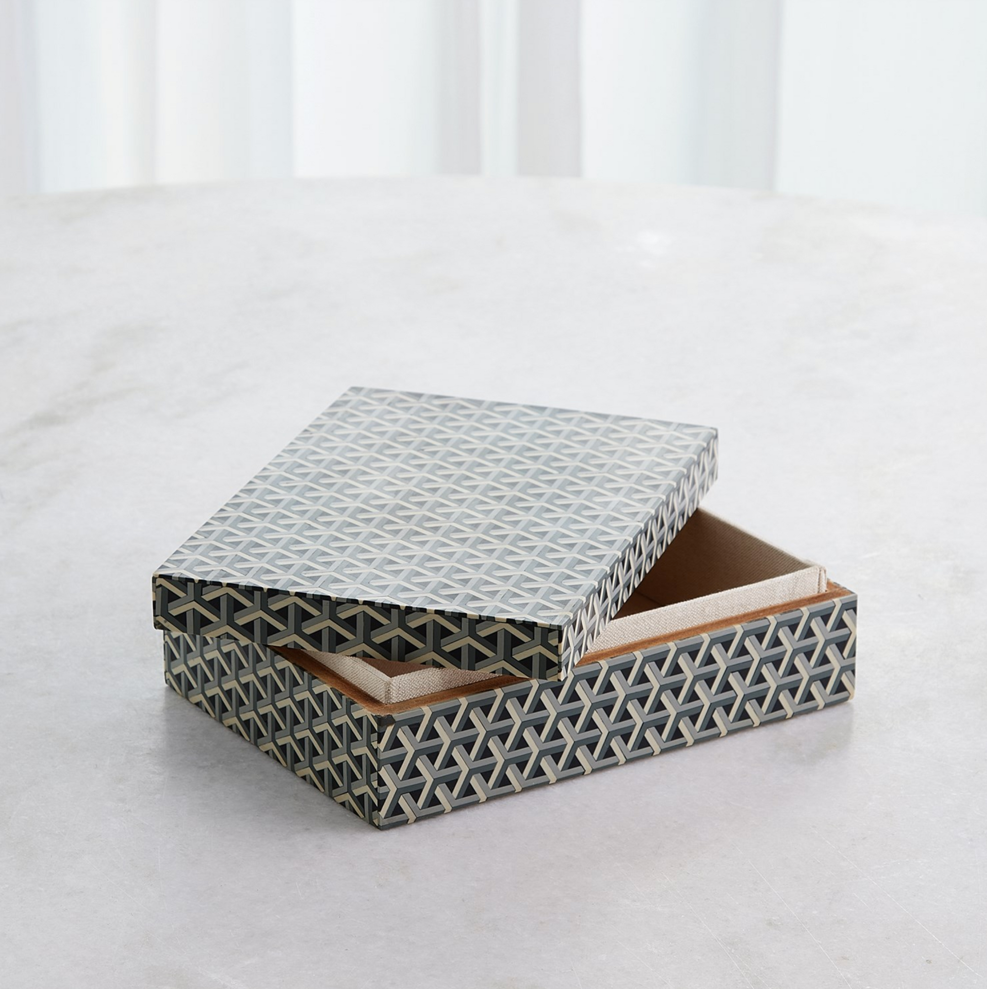 The Interwoven Box MD by Global Views is a decorative resin storage box featuring a blue and cream geometric pattern, pictured with its lid partially open on a light-colored surface.
