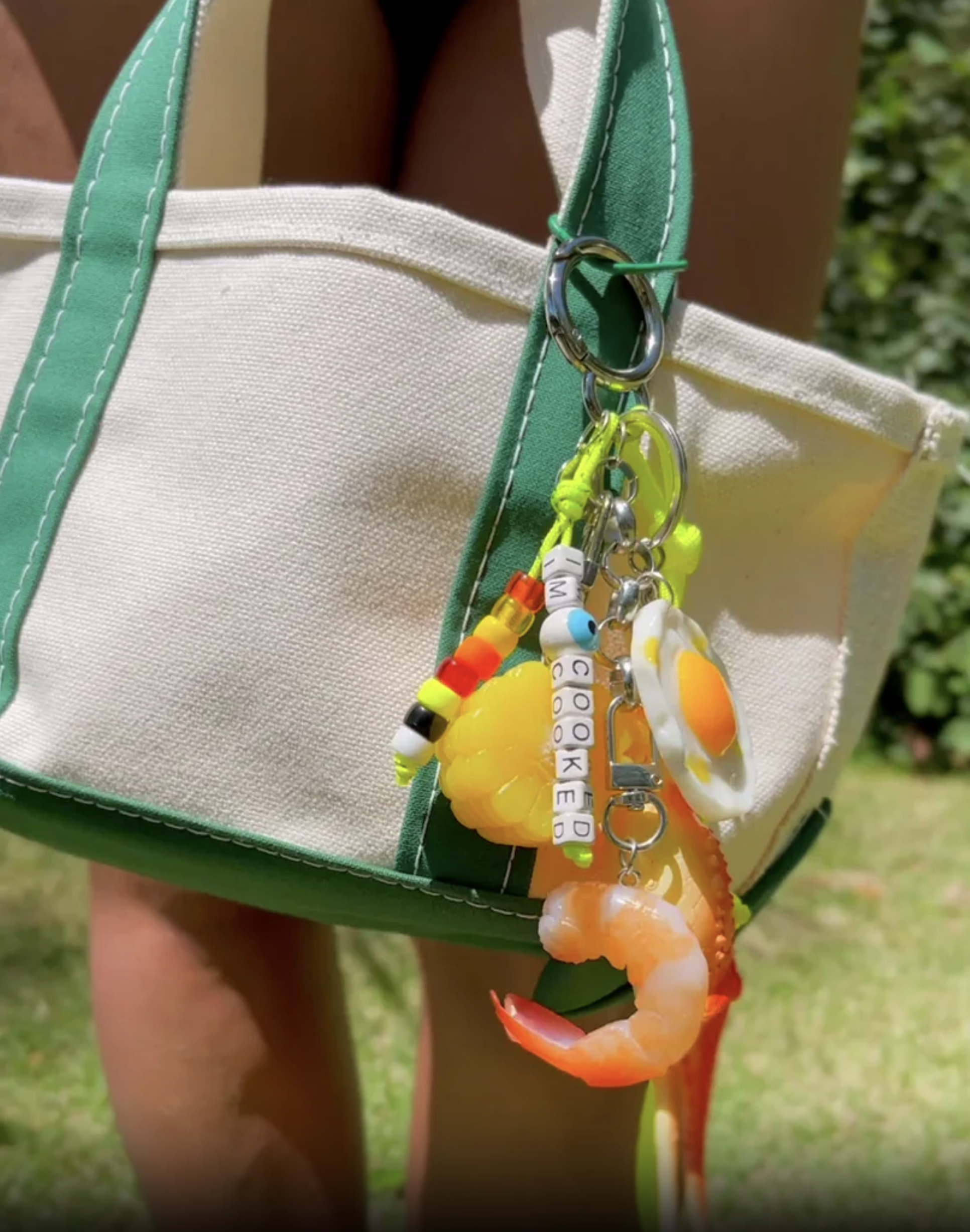 Outdoors, a Venessa Arizaga Bag Key Leash adorns a white and green tote bag, featuring food charms—plastic shrimp, fried egg, candy—and beads that spell &quot;I&#39;M COOKED,&quot; with grass and foliage in the background.