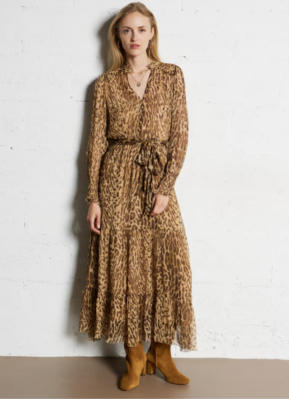 A woman with light hair stands against a white wall wearing the SUNCOO Cosmos Dress, a loose-fit, long brown leopard print style with a tie waist, paired with tan suede ankle boots and layered necklaces. Dress is 100% viscose.