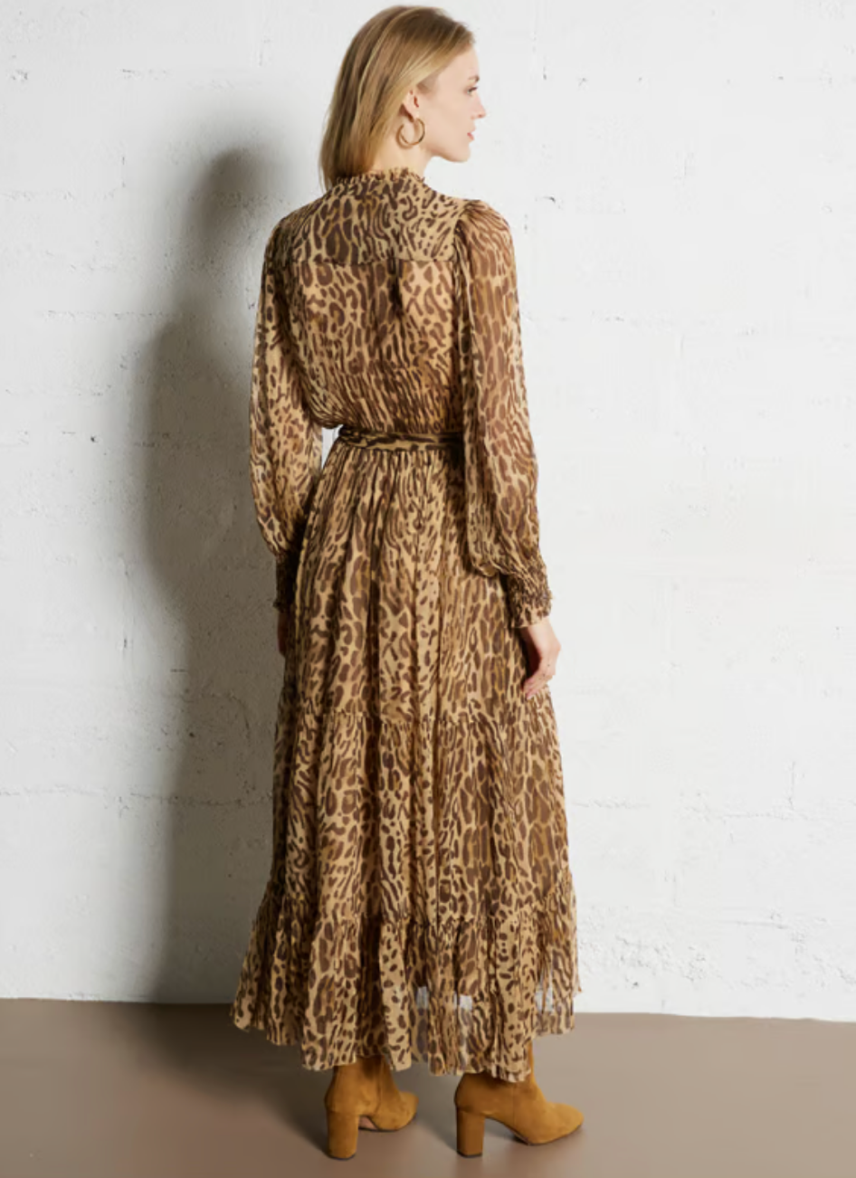 Wearing the SUNCOO Cosmos Dress, a woman faces away in a loose-fit, flowing leopard-print design with long sleeves and a thin belt. Made from 100% viscose, she styles it effortlessly with tan heeled ankle boots and gold hoop earrings.