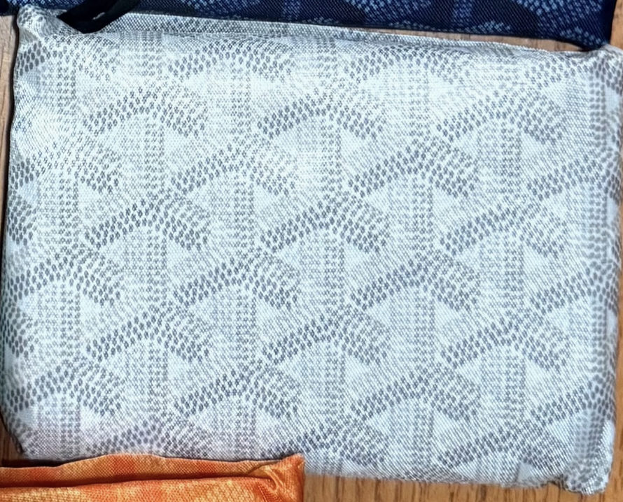 A close-up of Faire's Reusable Shopping Bags Assorted shows a designer-inspired fabric pouch with a gray and white geometric pattern on wood; an orange bag peeks from the bottom.