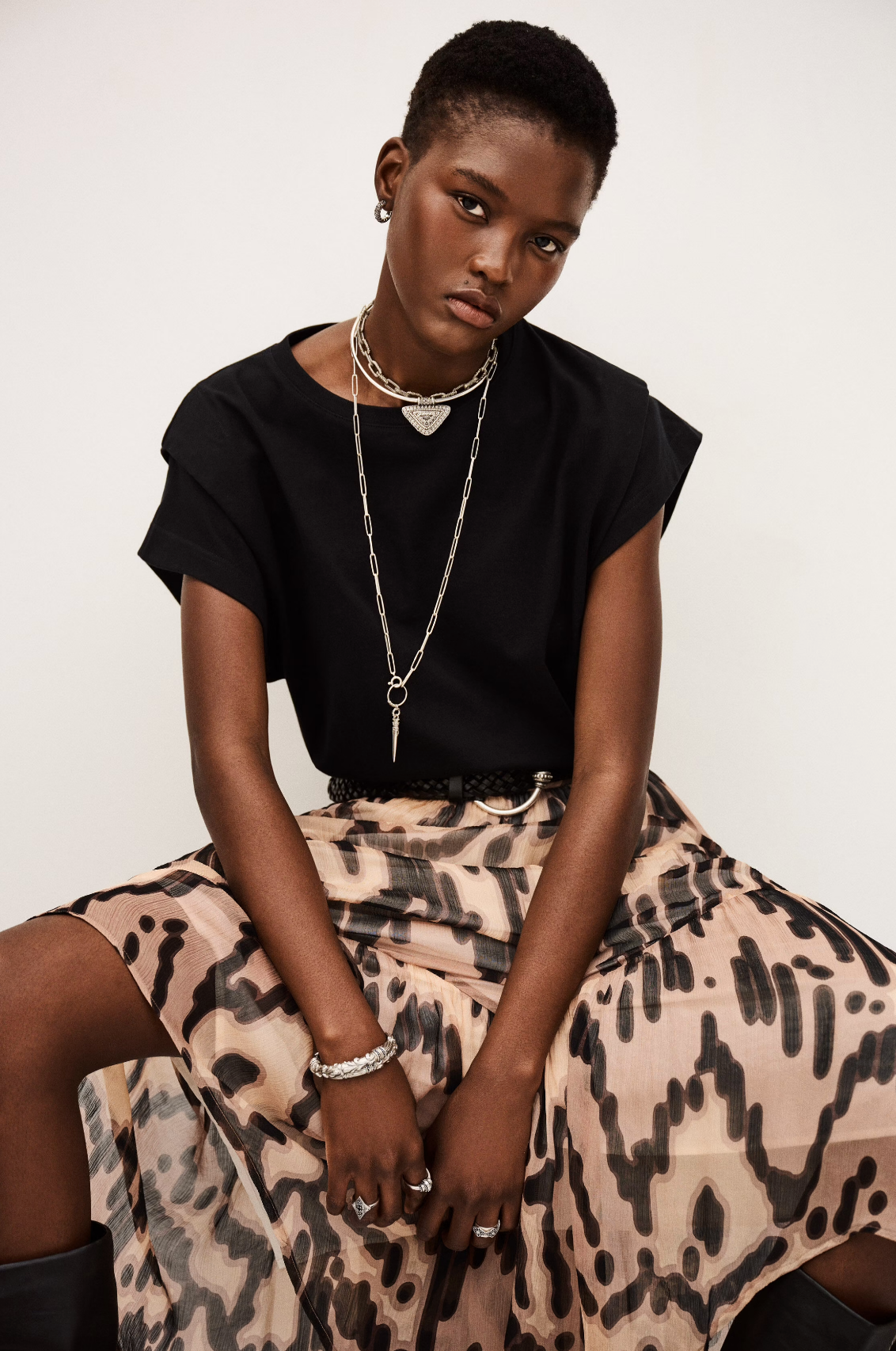 A person with short hair wears a black t-shirt, layered silver necklaces, and the BA&SH Skirt Aley, featuring a printed beige design. Seated with hands in their lap, they look at the camera with a neutral expression against a plain background.