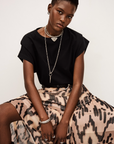 A person with short hair wears a black t-shirt, layered silver necklaces, and the BA&SH Skirt Aley, featuring a printed beige design. Seated with hands in their lap, they look at the camera with a neutral expression against a plain background.