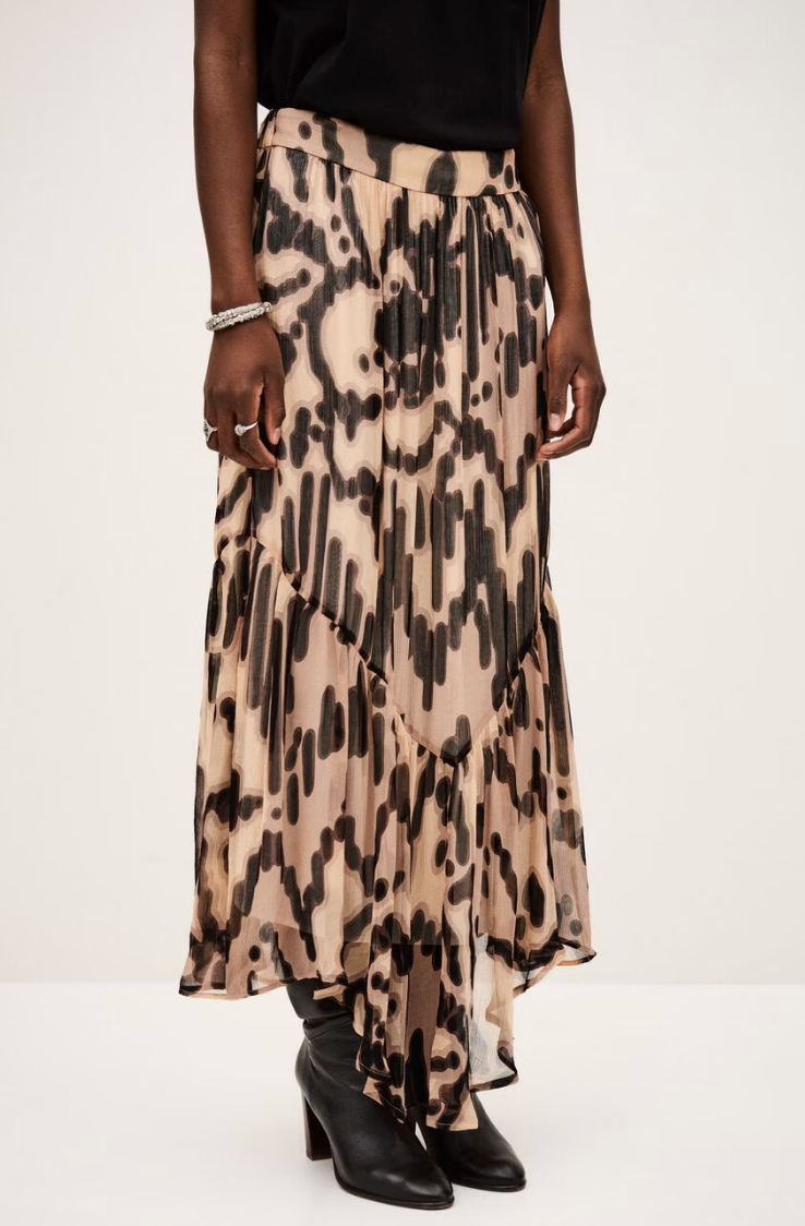 A person wearing the BA&SH Skirt Aley, a flowing beige maxi skirt with an abstract print, paired with black heeled boots, a black top, and a bracelet, stands against a plain light background.