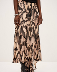 A person wearing the BA&SH Skirt Aley, a flowing beige maxi skirt with an abstract print, paired with black heeled boots, a black top, and a bracelet, stands against a plain light background.
