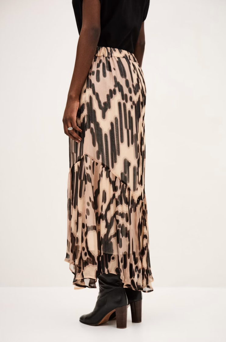 A person wears the BA&SH Skirt Aley—a flowy beige and black printed skirt with an asymmetrical cut—paired with a black top and black heeled boots, standing against a plain light background.