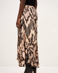A person wears the BA&SH Skirt Aley—a flowy beige and black printed skirt with an asymmetrical cut—paired with a black top and black heeled boots, standing against a plain light background.