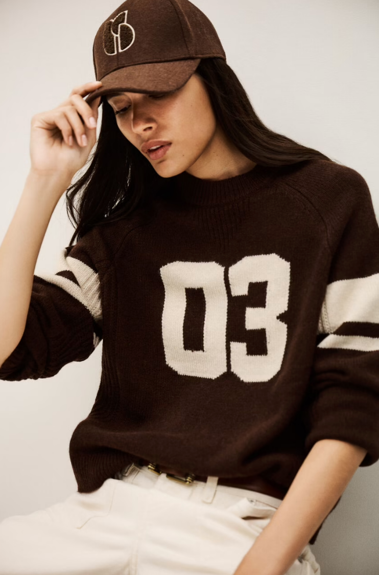 A person models the BA&SH Jumper Cobe, a brown cashmere sweater with "03" and white sleeve stripes, paired with a matching brown cap and light pants, posing with one hand holding the cap's brim.