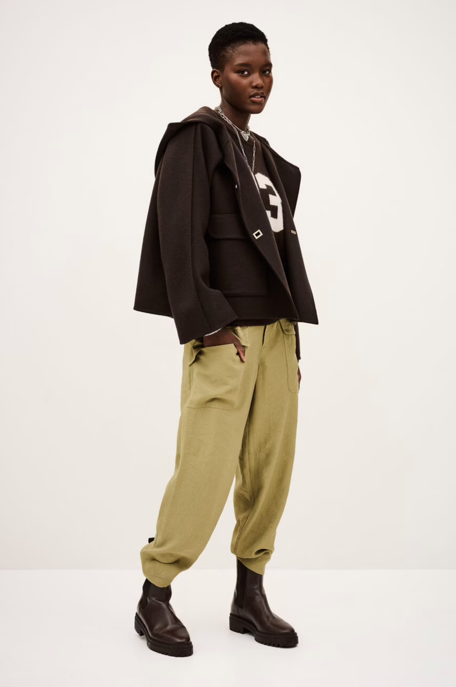 A person with short hair poses in the BA&SH Jacket Kove over a dark hoodie with a white “G,” light olive-green drawstring pants, and black boots, standing against a plain light background.