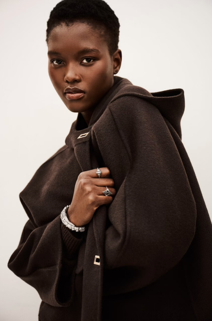A woman with short hair wears the BA&SH Jacket Kove, a dark brown, loose-fitting jacket with a hood, metallic accents, and wide collar. She holds it closed, showcasing her rings and bracelet as she looks confidently at the camera against a plain background.