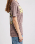 A woman with long curly hair stands in profile against a plain white background, wearing the DayDreamer Willie Nelson Red Headed Stranger Tee and blue jeans with large pockets.