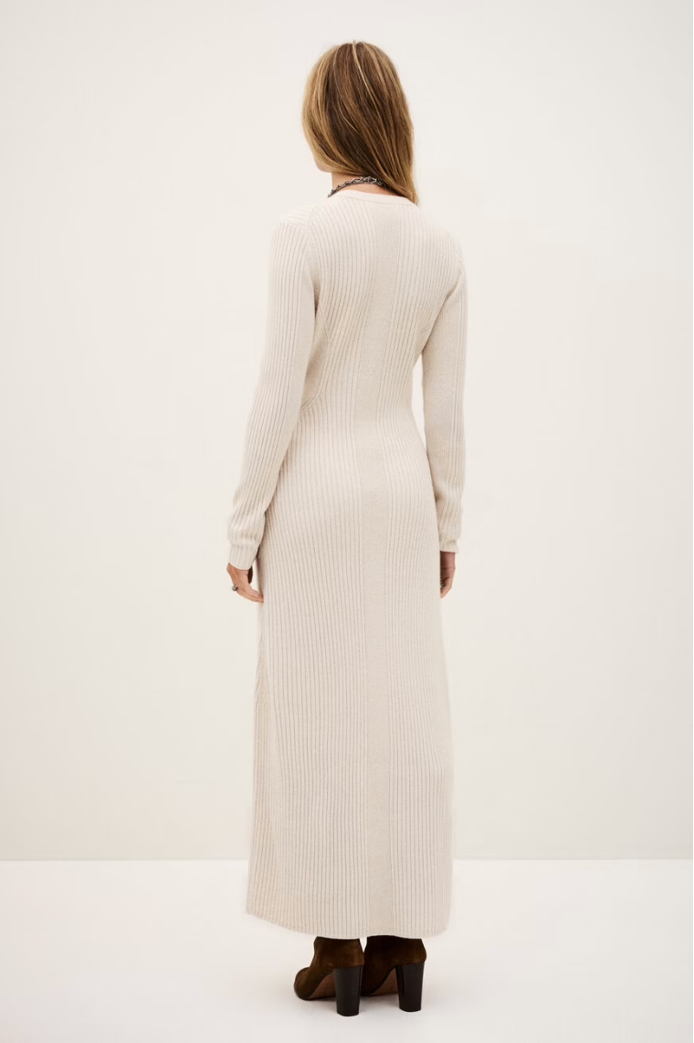 A woman with light brown hair wears the BA&SH Dress Anisa, a long, cream ribbed knit dress with long sleeves, made from a cozy polyester blend. She pairs it with brown heeled boots against a plain light background.