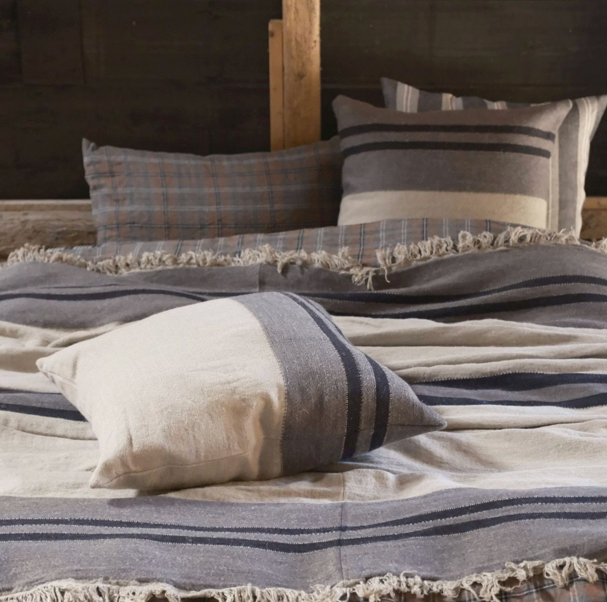 A cozy bed features Libeco Home’s Black House Stripe Pillow, styled with 25x25 inserts. The striped design in gray, cream, and navy rests against a wooden headboard, evoking rustic warmth and comfort.