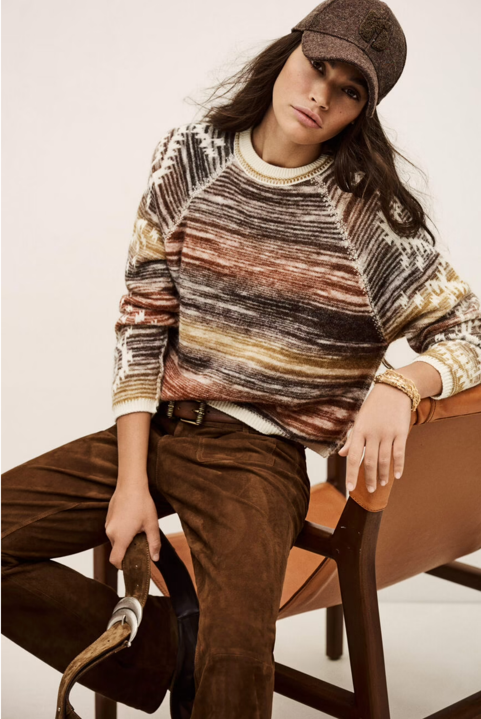 A person wearing the BA&SH Jumper Clodie, brown suede pants, and a brown cap sits on a tan chair holding a riding crop, looking relaxed and confident.