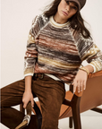 A person wearing the BA&SH Jumper Clodie, brown suede pants, and a brown cap sits on a tan chair holding a riding crop, looking relaxed and confident.