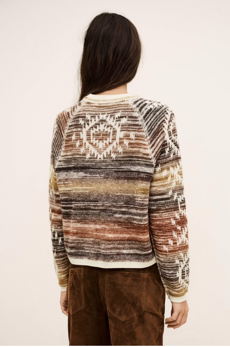A person with long dark hair, seen from behind, is wearing the BA&SH Jumper Clodie—a patterned, multicolored alpaca wool sweater in earth tones—paired with brown corduroy pants against a plain light background.