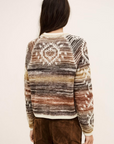 A person with long dark hair, seen from behind, is wearing the BA&SH Jumper Clodie—a patterned, multicolored alpaca wool sweater in earth tones—paired with brown corduroy pants against a plain light background.