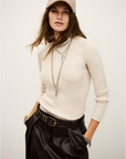 A woman poses with her hands in her pockets wearing the BA&SH Jumper Banny, layered necklaces, a beige baseball cap, black belted trousers, and bracelets against a plain background.
