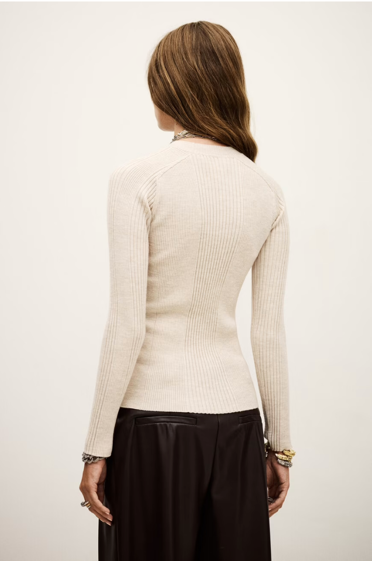 A woman with long, light brown hair is shown from the back wearing the BA&SH Jumper Banny, a fitted, ribbed, cream-colored sweater with long sleeves, paired with a black skirt. She has bracelets on her wrist and stands against a light background.