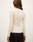 A woman with long, light brown hair is shown from the back wearing the BA&SH Jumper Banny, a fitted, ribbed, cream-colored sweater with long sleeves, paired with a black skirt. She has bracelets on her wrist and stands against a light background.